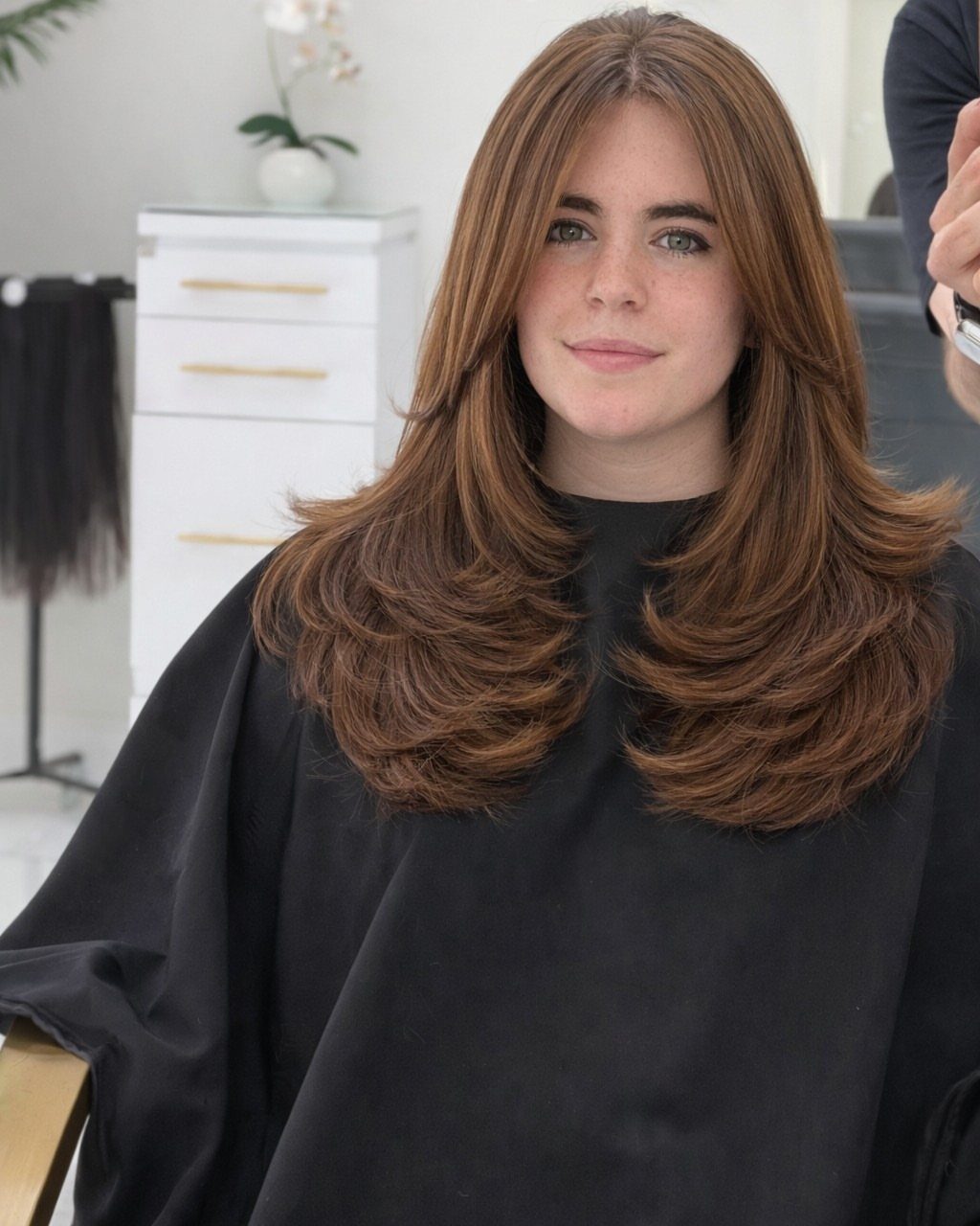 Woman with chestnut-brown face-framing layers and curled ends in a salon chair