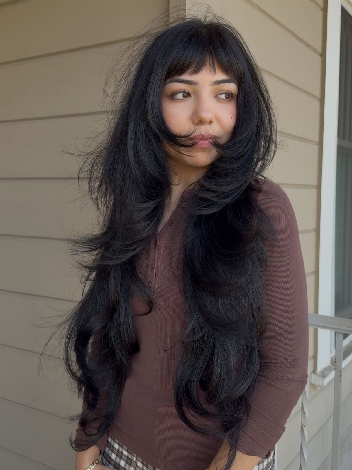 Woman with long layered black hair and short wispy bangs standing by beige siding