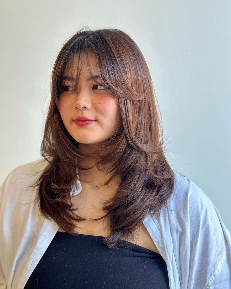 Woman with a round face wearing long layered brown hair and curtain bangs